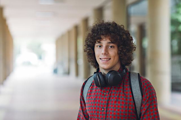 A young male student smiling with headphones around his neck and a backpack on. 